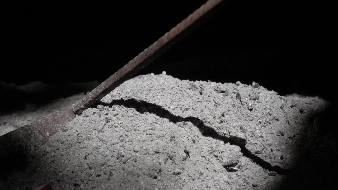 Metal tool rakes pile of ash in dimly lit indoor setting, showing fine debris texture