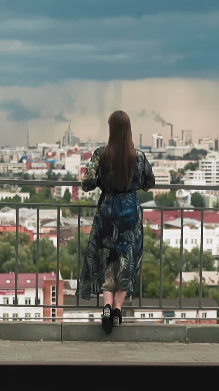 Brunette lady with long hair wearing elegant dress with colorful decor leans on handrail looking at city from viewing deck slow motion backside view