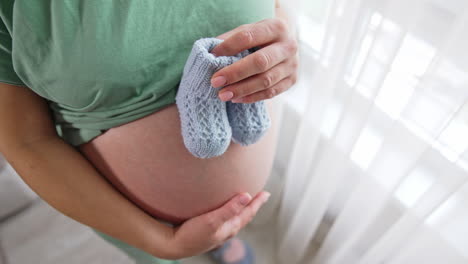 pregnant lady holding two cute baby booties over her belly. she puts booties on her belly and steps with them slowly over her tummy. view from the above.