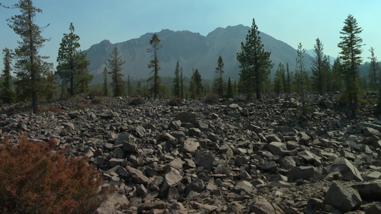 Kivinen vulkaaninen maasto hajallaan olevilla havupuilla ja vuorijono taustalla, Lassen Volcanic National Park, Kalifornia, USA. Uloszoomauskuva.