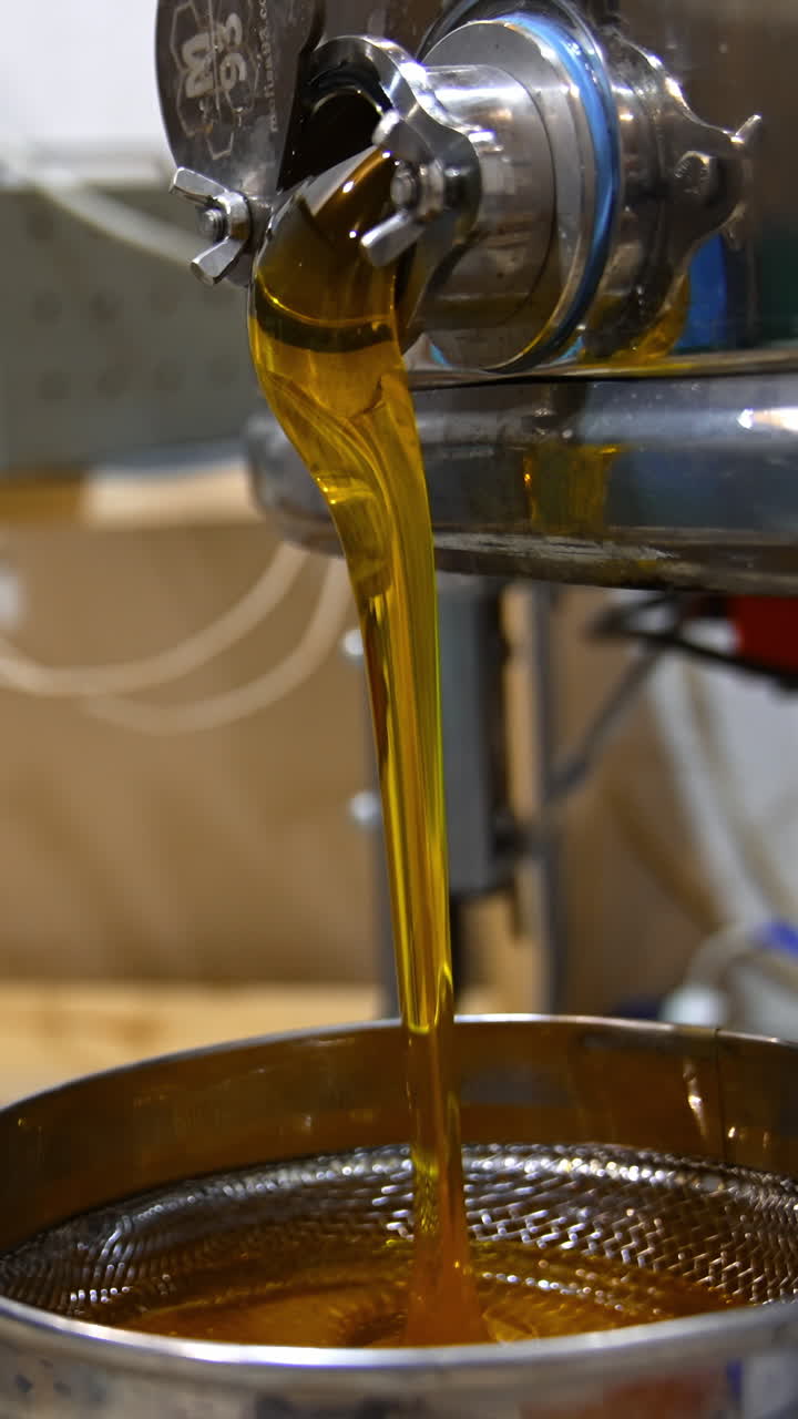 Fresh amber honey quickly flowing from metal barrel faucet. Organic product being poured onto sieve for filtration. Blurred backdrop. Vertical video