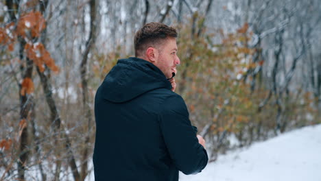 following a man walking by the park holding a phone at his ear. man with coffee cup stops and turns to camera speaking. winter nature at backdrop.