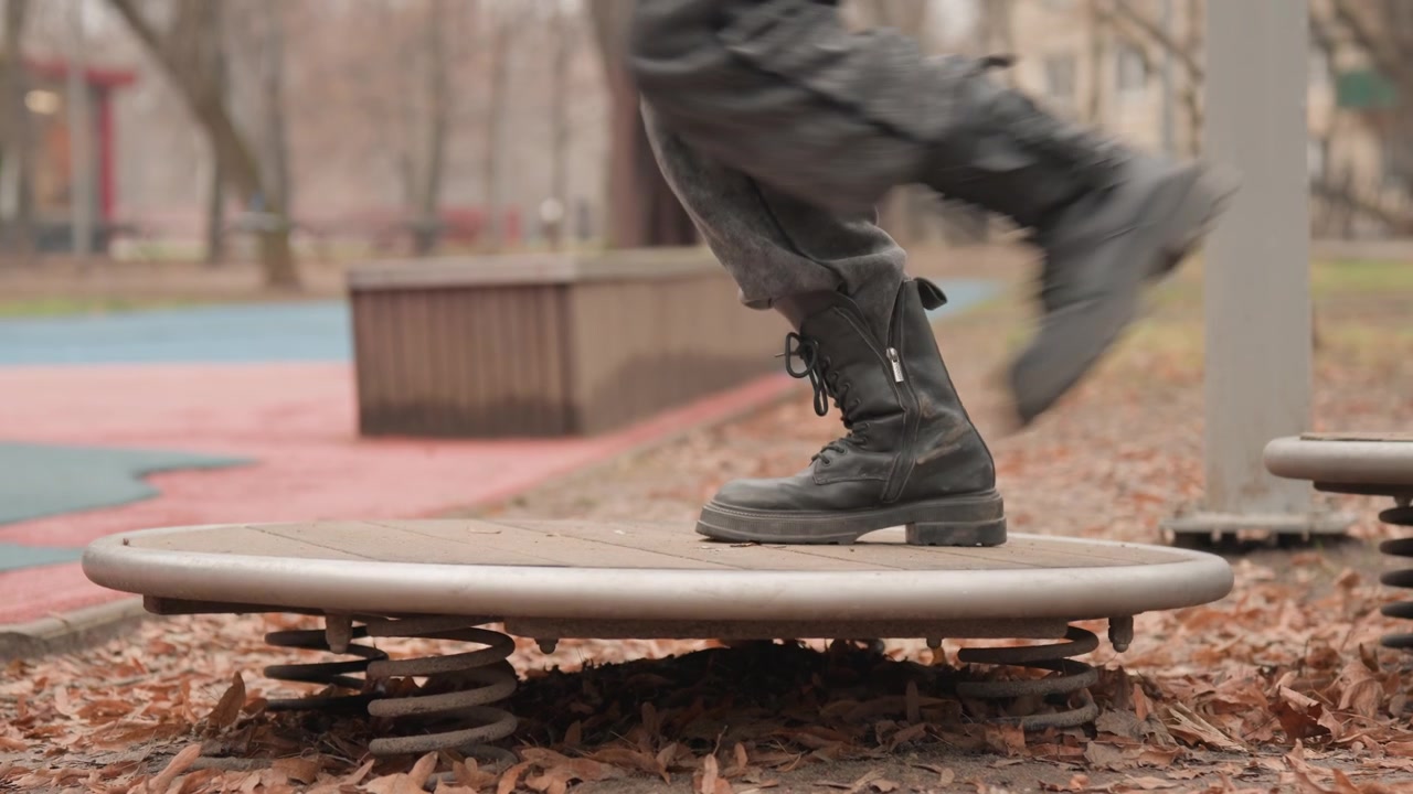 Single boot hopping on round spring platform, playground coil compresses and rebounds while person explores motion and rhythm, vibrant yet muted urban park setting with benches and onlookers blurred