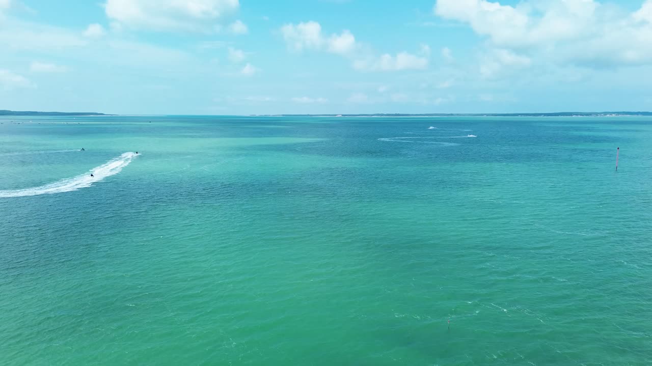 A dynamic drone footage captures people riding jetskis in the waters of Tremblade, Charente, France, creating trails of white foam against the blue sea on a sunny day
