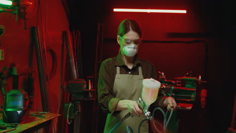 caucasian woman in goggles, respiratory mask and apron working with pulverizator in workshop