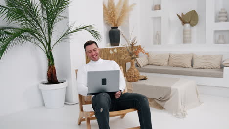positive caucasian male wearing white sweater and jeans sits in armchair. man laughs cheerfully holding a laptop in hands.