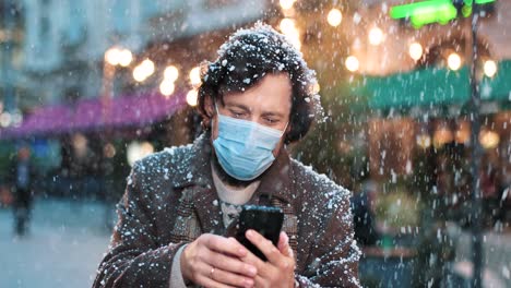 close-up view of caucasian man wearing a face mask and using smartphone on the street while it‚äôs snowing in christmas