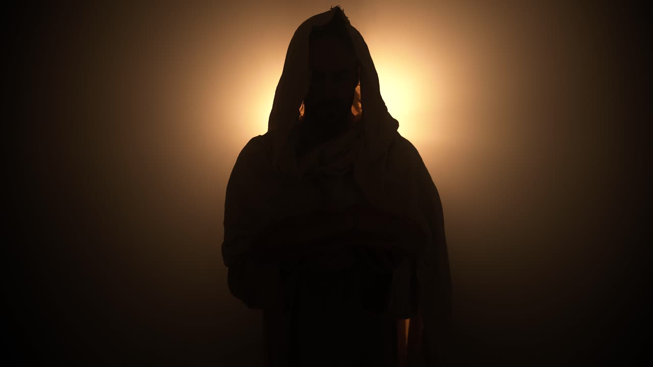 Silhouette of Jesus Christ in white hooded robe holding bread as he stands in glowing, bright light praying. Perfect for representation of Jesus as the bread of life, last supper, communion.
