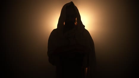 silhouette of jesus christ in white hooded robe holding bread as he stands in glowing, bright light praying. perfect for representation of jesus as the bread of life, last supper, communion.