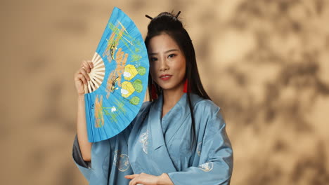 junge asiatische frau im blauen kimono winkt mit einem fan und versteckt ihr gesicht, während sie lächelt und flirtet