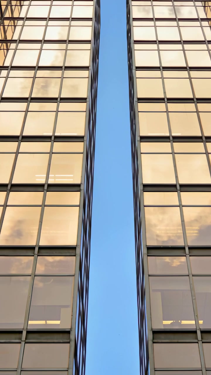 Video verticale: Telecamera inclinata che mostra uno stretto squarcio di cielo blu in un canyon urbano incorniciato da facciate di vetro.