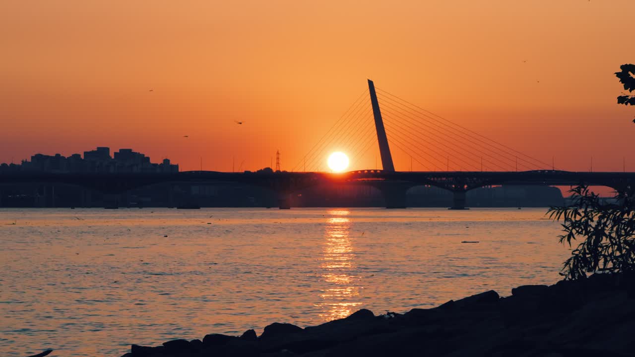 Sun setting behind the silhouettes of the Seongsan and World Cup Bridges spanning the Han River in Seoul, South Korea, with a golden reflection on the water and an orange sky