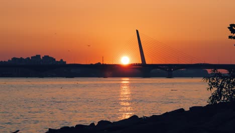 sun setting behind the silhouettes of the seongsan and world cup bridges spanning the han river in seoul, south korea, with a golden reflection on the water and an orange sky