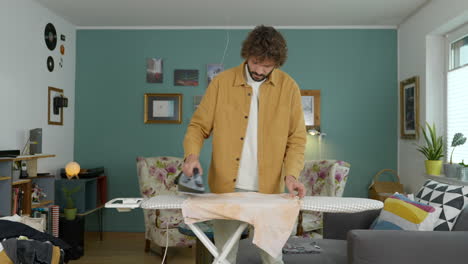 modern caucasian man ironing clothes in stylish living room doing housekeeping chores