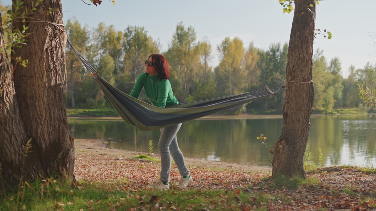 Donna bianca appende un'amaca in riva al lago, regolando le cinghie tra gli alberi, maglione verde e occhiali da sole, luce autunnale dorata che si riflette sull'acqua, postura rilassata mentre l'amaca si sistema, ritiro solitario e tranquillo
