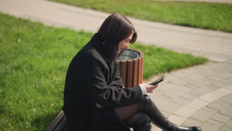 side view of young lady in black coat and knee-high boots sitting on a park bench, focused on smartphone, enjoying sunny day outdoors with greenery surrounding