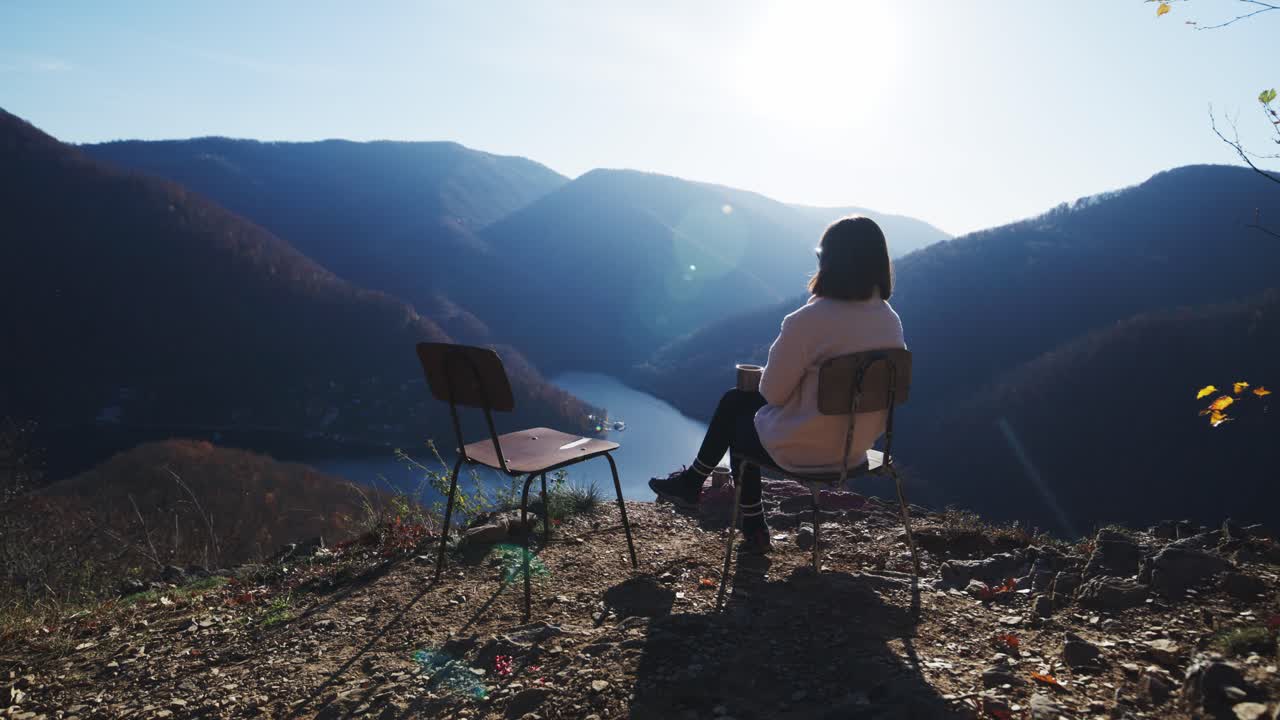 Medium shot of a woman sitting on a chair on a mountain ridge, contemplating the river gorge below. Bright sun flare adds a lens effect, emphasizing freedom, tranquility, and travel