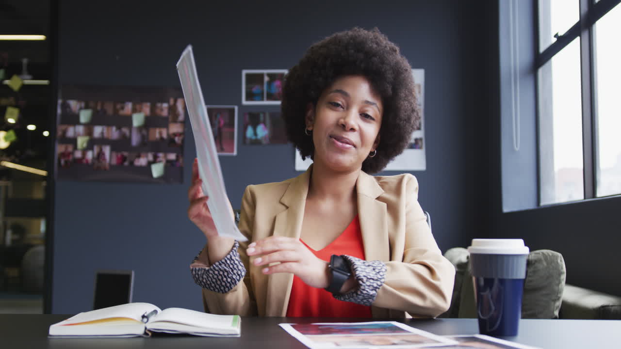 mujer de negocios biracial sentada teniendo un chat de video pasando por el papeleo en una oficina moderna