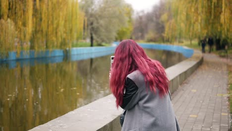 Mujer-Joven-Y-Atractiva-Con-Pelo-Rojo-Tomando-Fotos-En-Su-Teléfono-Inteligente-Mientras-Está-De-Pie-Junto-A-Un-Estanque-Artificial-En-Un-Parque-De-Otoño