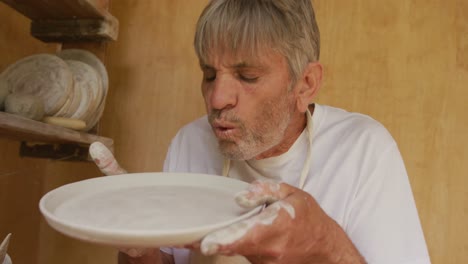 hombre caucásico mayor que usa un delantal inspeccionando un plato de arcilla en un taller de cerámica