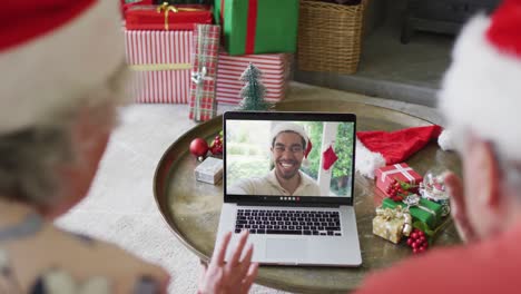 Älteres-Kaukasisches-Paar-Mit-Weihnachtsmützen-Nutzt-Laptop-Für-Weihnachtsvideoanruf-Mit-Mann-Auf-Dem-Bildschirm