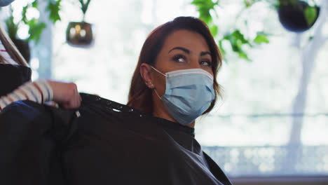 Female-hairdresser-putting-cape-on-female-customer-wearing-face-mask-at-hair-salon