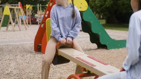 una niña con síndrome de down y su amiga rubia balanceándose en una mecedora de madera en el parque en un día ventoso