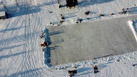 winter aerial rise twist over residential detached homes at empty manmade ice rink connected to skating track double oval of where people practice skating at community roadside park hay stack cube 2-3