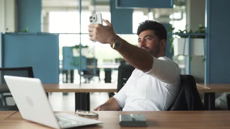 portrait of a handsome, stylish young man of arab descent sitting in a modern business center office with a laptop, taking a selfie on a smartphone