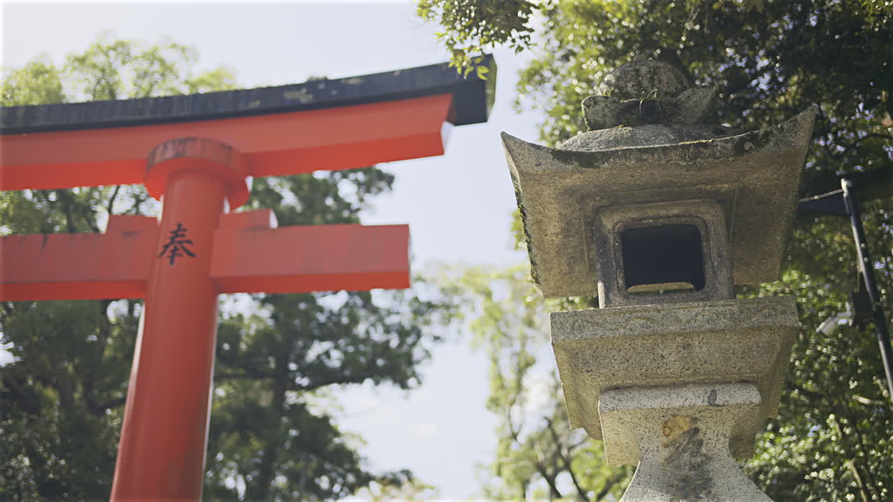 A vibrant red torii gate stands tall against a clear sky, while a stone lantern rests nearby, showcasing traditional architecture. Fushimi Inari, Kyoto, Japan