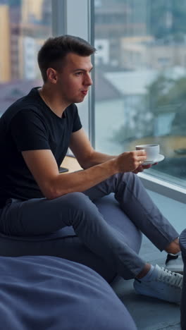 caucasian man sits on the chair drinking coffee. male employee looks into window relaxing from work at computer. blurred backdrop. vertical video