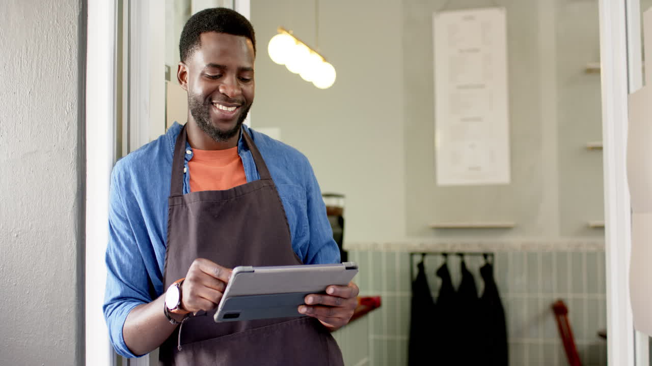 Bariste homme afro-américain avec un sourire chaleureux, tenant une tablette dans un café.
