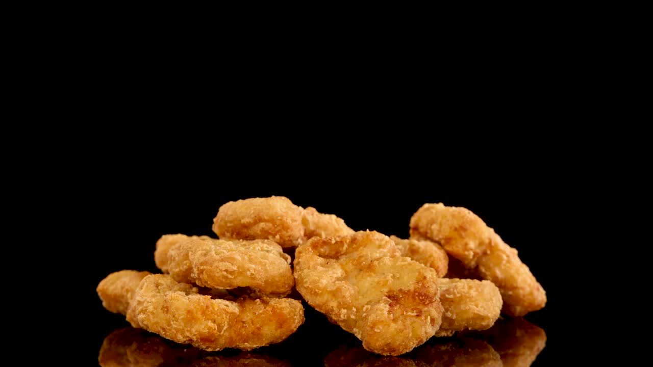 A hand reaches in to pick up golden, crumbed chicken nuggets from a pile on a glossy black surface, under bright studio lighting with smooth camera movement