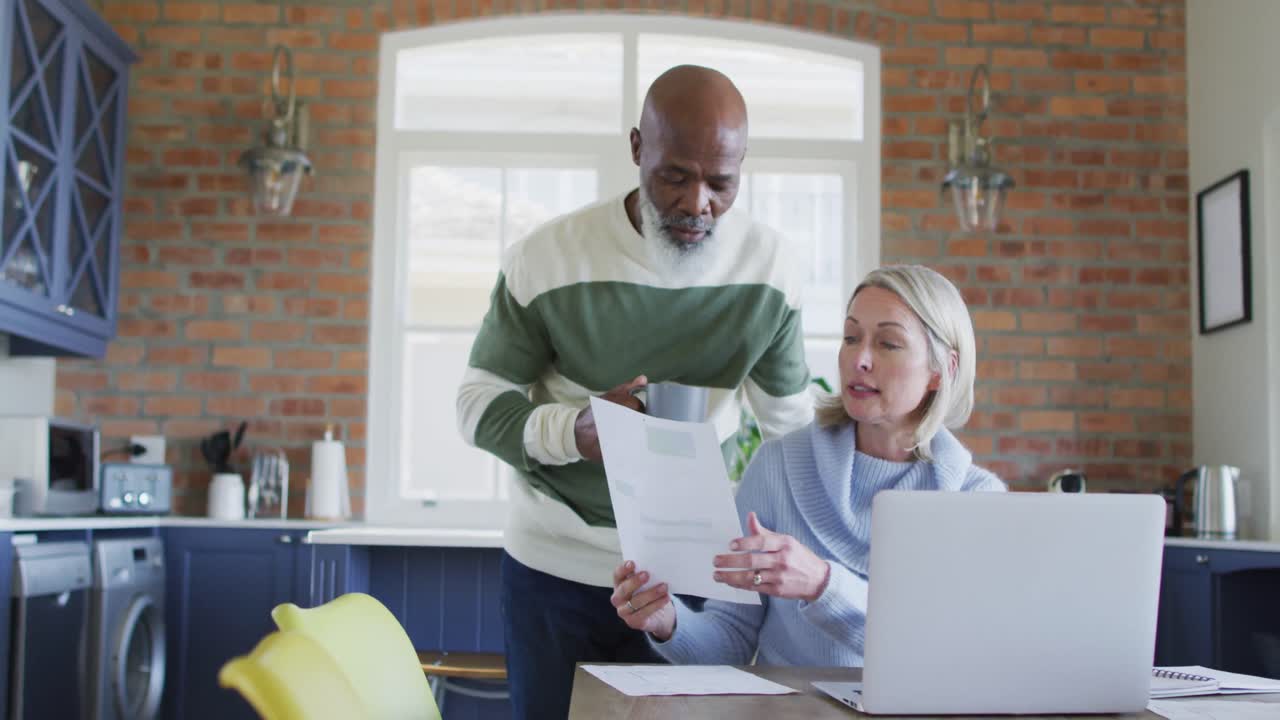 Stressed senior diverse couple in kitchen sitting at table, using laptop