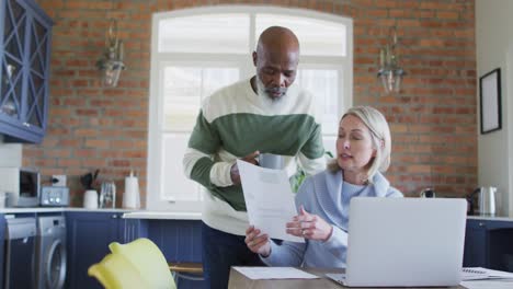 stressed senior diverse couple in kitchen sitting at table, using laptop