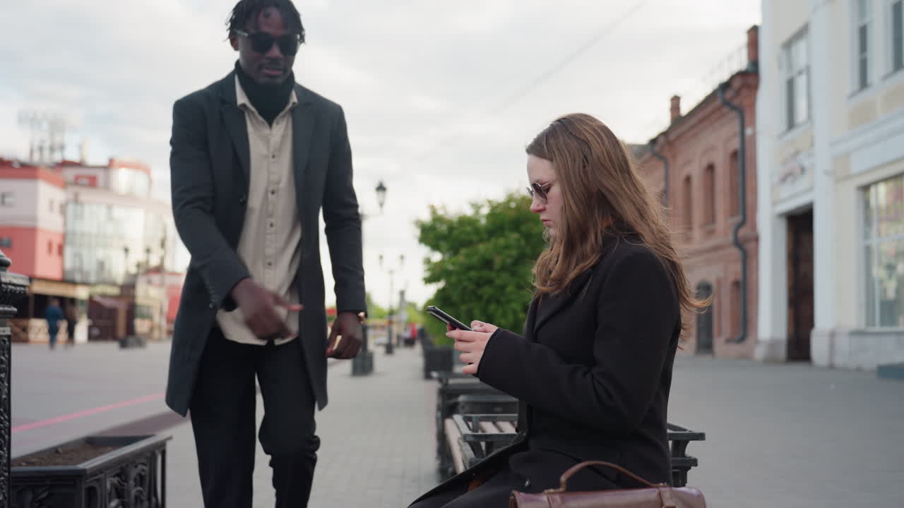 Man walks past woman using phone on city bench while cars move in distant background, surrounded by red buildings, urban streetlights, and soft evening sky creating modern calmness and vibe