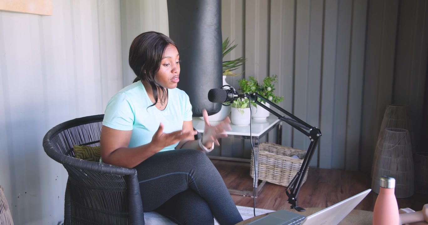 Afroamerikansk kvinde taler ind i en mikrofon, henviser til en bærbar computer og gestikulerer, mens hun podcaster derhjemme.