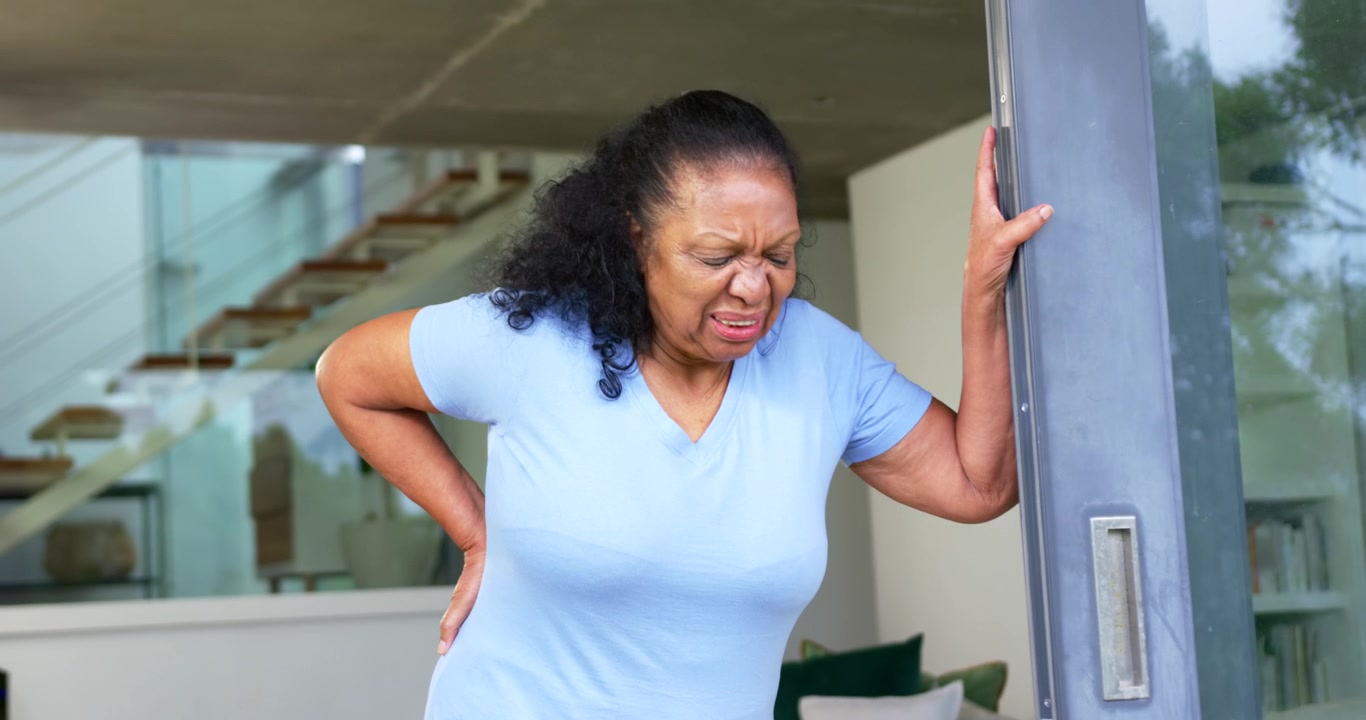 Senior African American woman wincing from back pain, leaning on sliding door at home, steadying