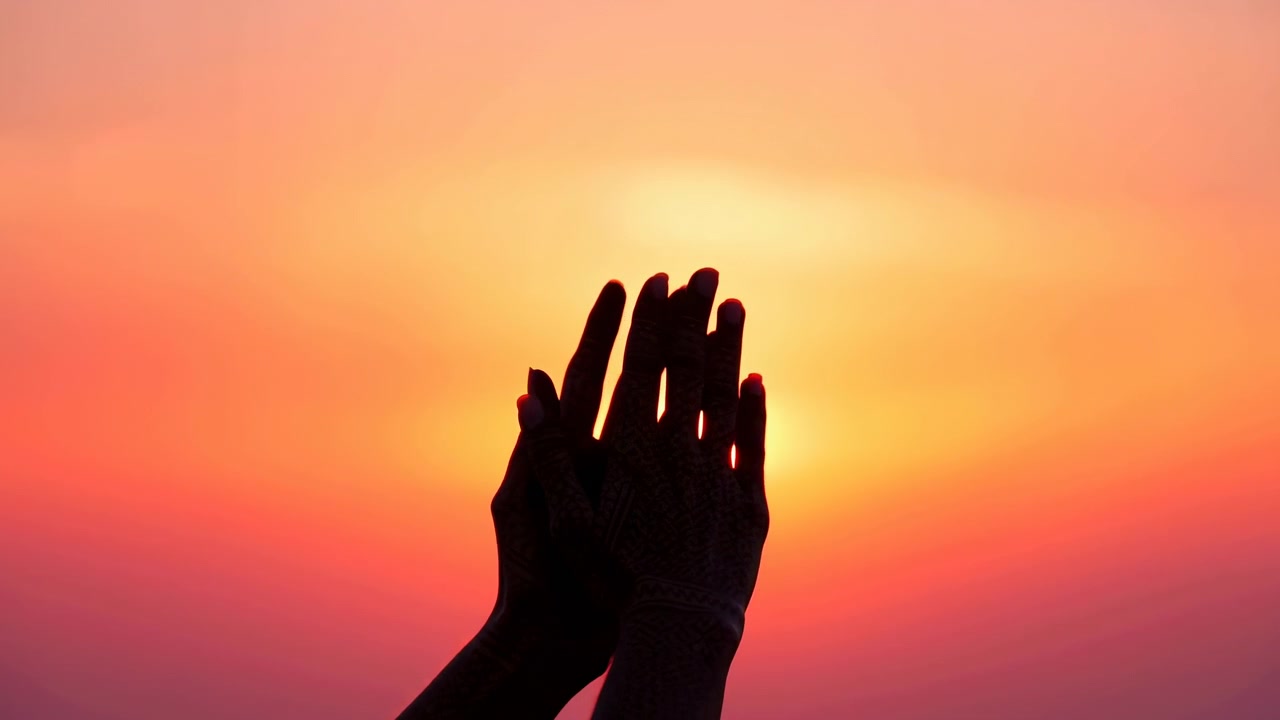 Due mani in silhouette si alzano dalla parte inferiore dell'inquadratura e si aprono delicatamente con le dita estese verso il sole splendente dell'alba, creando un gesto di offerta riverente contro un vasto cielo sfumato e un'atmosfera calda e retroilluminata.