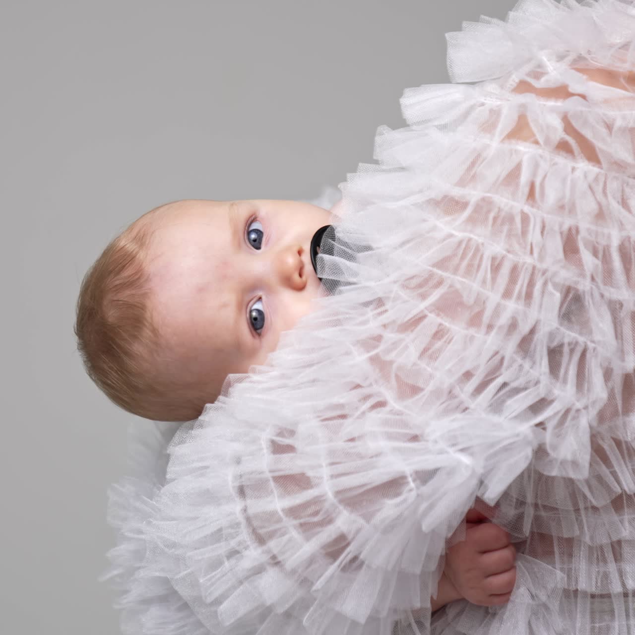 Unrecognized long-haired blond woman holding a baby. Tiny infant with pacifier looking at camera. White backdrop