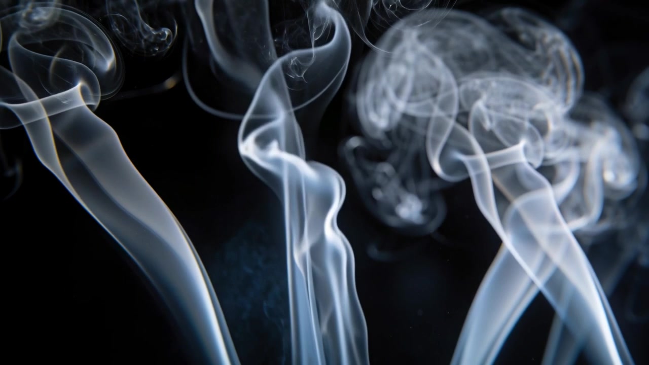 Pennacchi di fumo che si alzano, si arricciano e si fondono da sotto l'inquadratura in uno studio buio, rivelando un bagliore illuminato dal bordo.