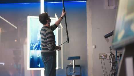 man holding boom microphone, adjusting it in modern studio with neon lights, equipment, and minimalistic background, focusing on sound recording during professional video production process