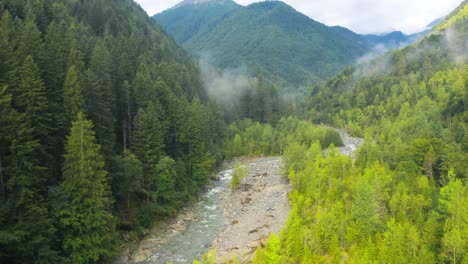 Drone-Flight-along-a-small-river-in-a-valley-of-Spruga,-Locarno-alps-Mountains,-between-green-forests,-slow-motion,-mountains-all-around,-Switzerland,-italy