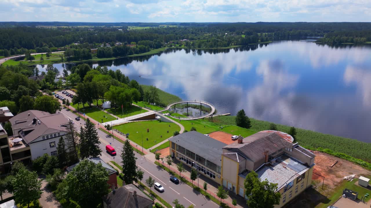 Wide drone shot showing the town of Zarasai along a reflective lake, with rooftops, streets, and forested hills. Shot at Zarasai, Lithuania (Zarasai, Lietuva)