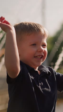 smiling little boy waves hand splashing fountain water on mother in sunny park closeup slow motion. happy childhood. cute child plays near fountain