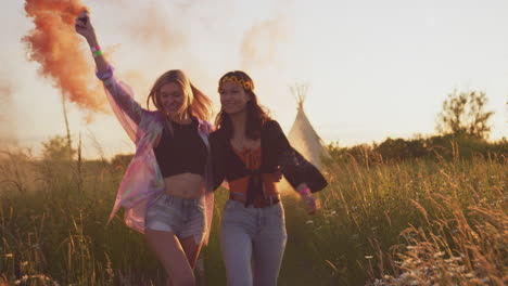 two female friends camping at music festival running through field with smoke flares