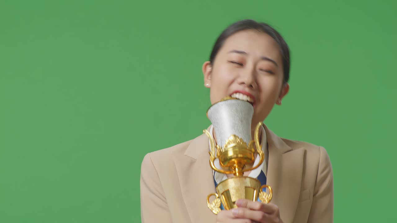 금메달을  슈트를 입은 아시아 사업가 여성이 골드 트로피에서 노래하고 스튜디오의 녹색 스크린 배경에서 첫 번째 우승자로 우승을 축하합니다.