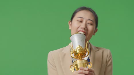 close up of asian business woman in a suit with a gold medal singing on a gold trophy celebrating winning as the first winner on green screen background in the studio