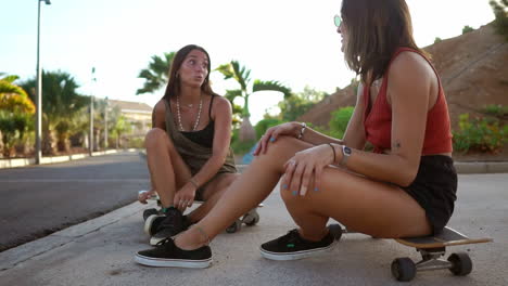 dos chicas se sientan en sus patinetas en el parque de patinaje durante la puesta de sol, inmersas en una conversación, sus sonrisas y risas reflejan la camaradería de los amigos. una conversación relajada de longboard