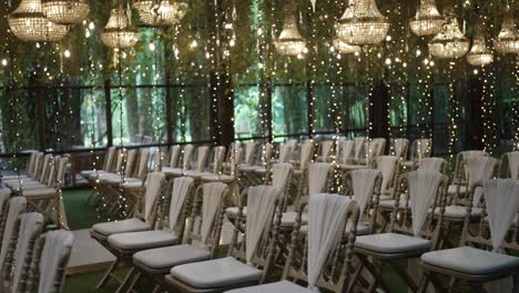 rows of white chairs with soft fabric decor beneath glowing string lights and chandeliers